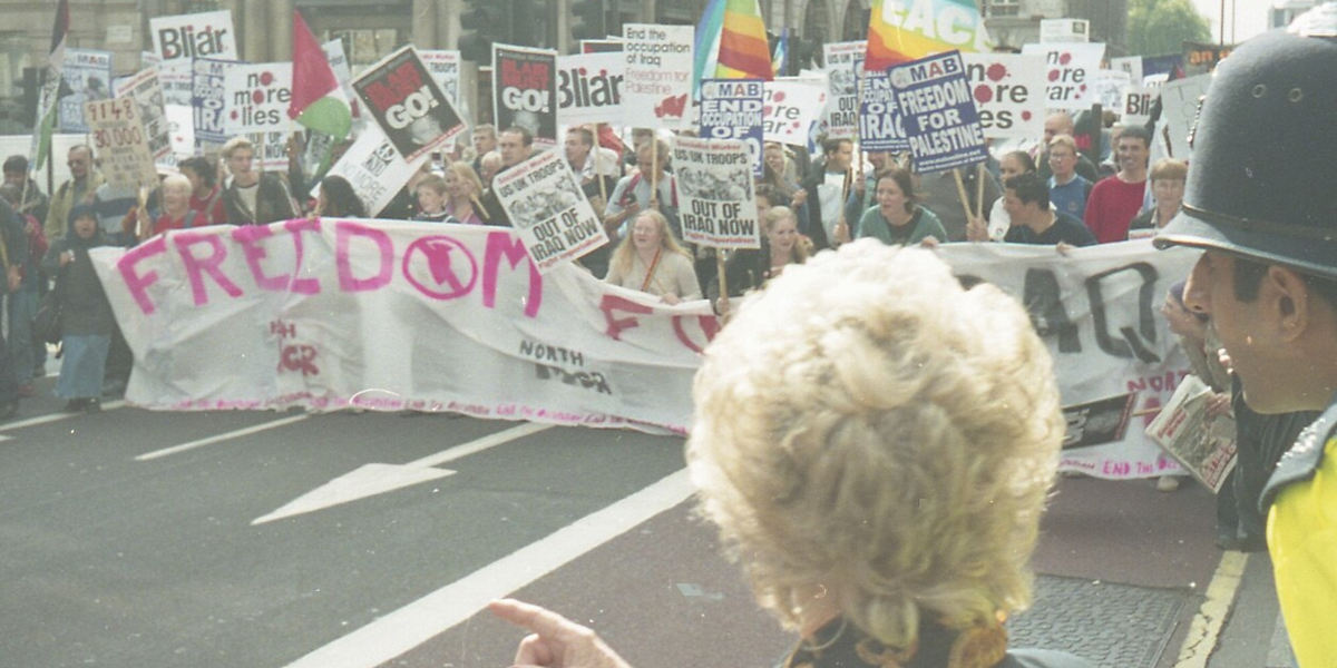 Protestors waiving placards denouncing the Iraq War and marching behind a banner. A woman and a policeman are in the foreground