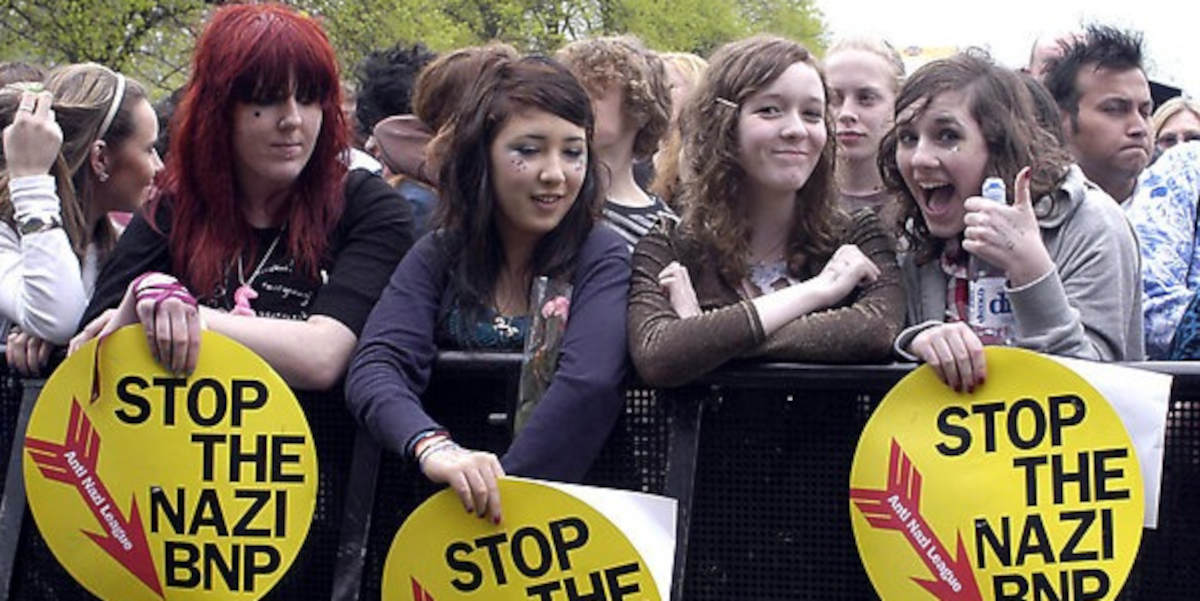Young women festival-goers stand at a gig barrier holding placards that read 'Stop the Nazi BNP' while smiling and putting thumbs upCREDIT: PA PHOTOS