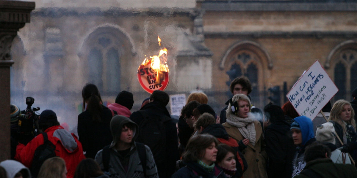 Student protestors in London in 2010, with one holding a burning placard reading 'Stop fees and cuts'