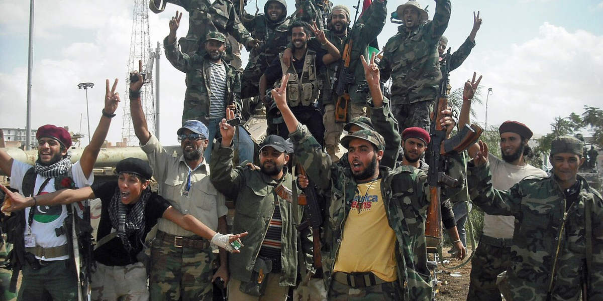 A group of armed men, standing around a truck, celebrating and throwing up victory signs