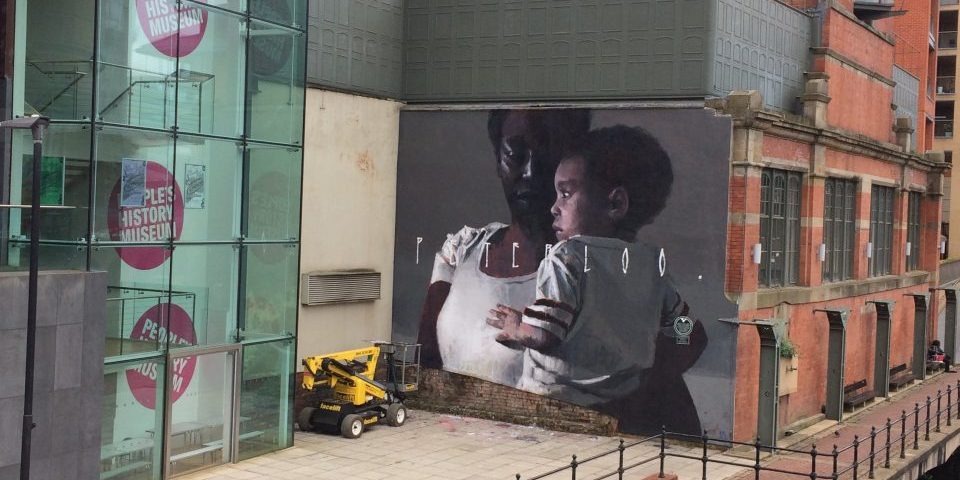 The outside of a glass-fronted building next to a wall showing a large painting of a black woman holding a baby. both wearing white t-shirts, with the word 'Peterloo' written across