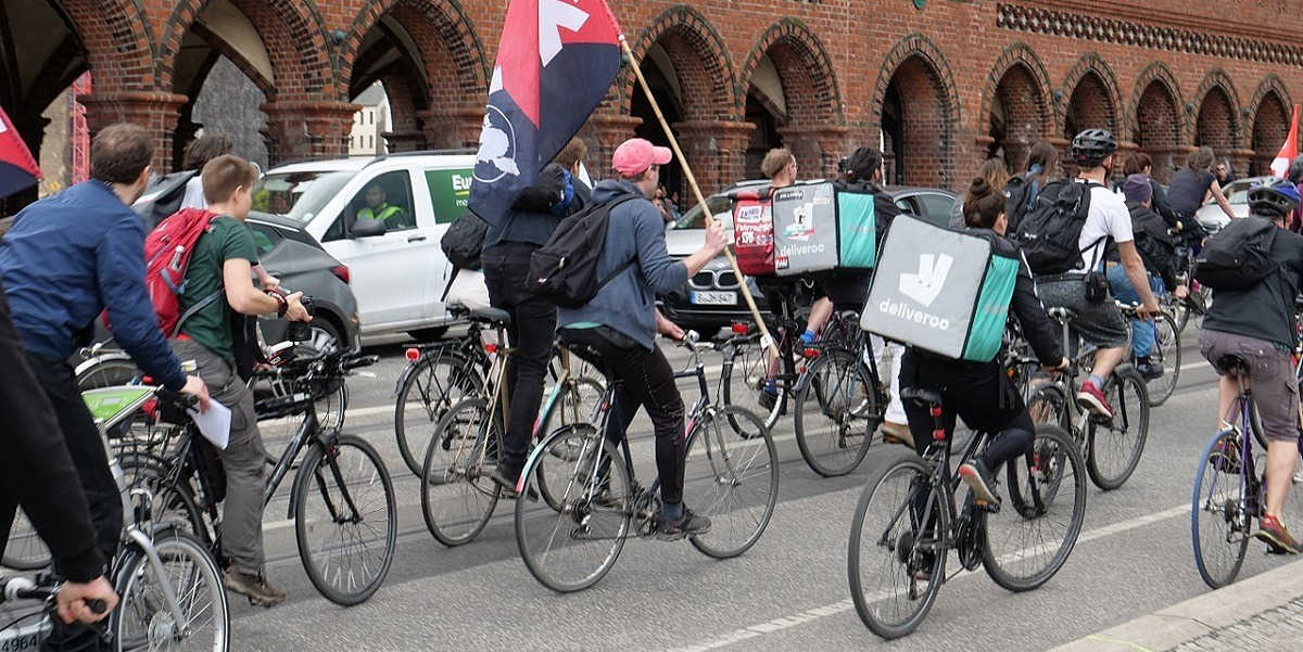 A group of cyclists with Deliveroo bags on their backs and holding protest flags travel in convoy during the day in Berlin