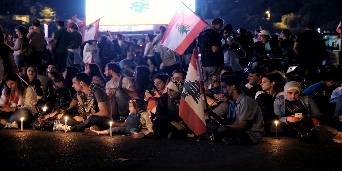 A crowd of people with Lebanese flags and candles sit and stand in a group at night