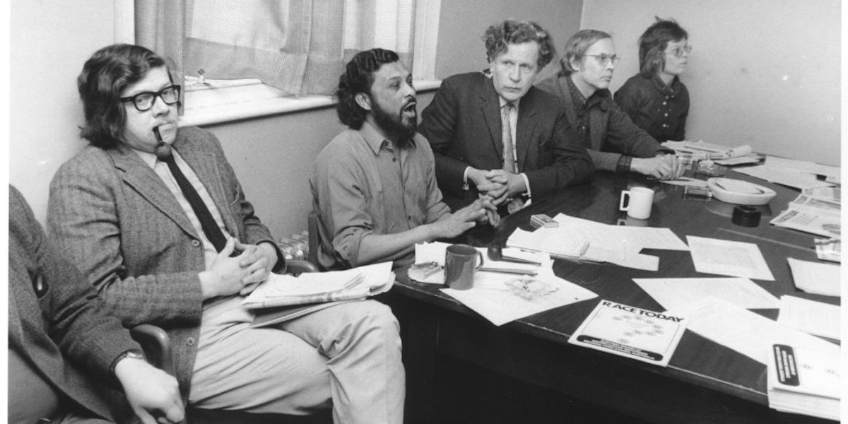 A black and white photograph shows a panel of speakers sitting behind a table in the 1970s