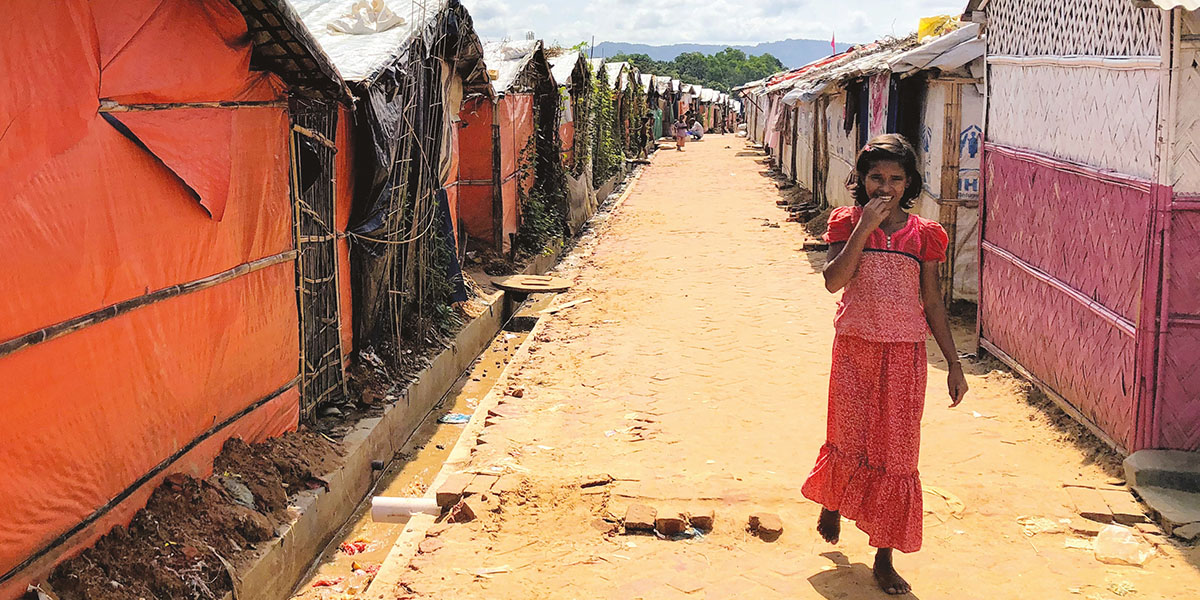 A young girl in a red skirt and top walks along a sandy road lined by shacks, in a refugee camp
