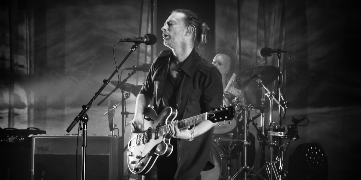 Black and white photograph of Thom Yorke singing and playing guitar with band in the background