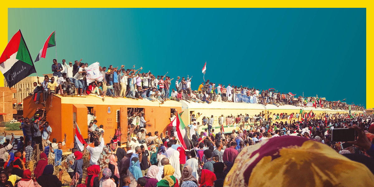 Photo of a long train covered and surrounded by hundreds of people wearing bring colours and waving Sudan flags