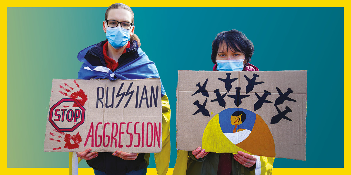 Two white feminine people in masks, their shoulders draped in Ukraine flags, hold cardboard signs. One reads: Stop Russian aggression, the other shows a drawing of bombs falling on a mother and child