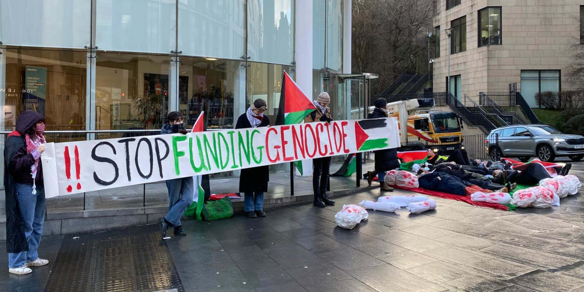 A protest outside a glass-fronted building includes people lying on the floor in a die-in pose next to five people holding a banner that reads: !! STOP FUNDING GENOCIDE with a flag of Palestine