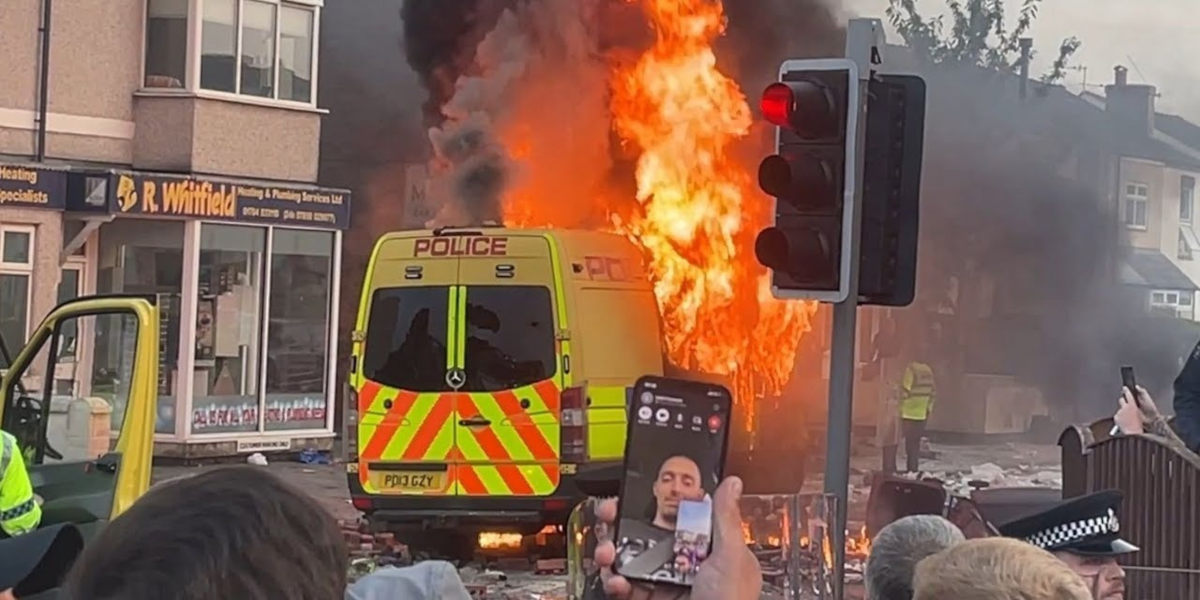 A police van on fire in a English town high street. In the foreground, in front of traffic lights are heads and hands from a crowd of people filming the scene, or themselves speaking to cameras