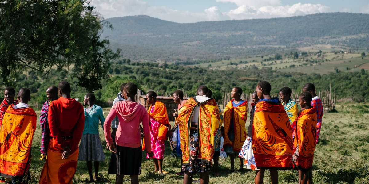 Against a background of green fields and mountains, a group of people wearing primarily traditional Maasi clothes stand in a circle talking