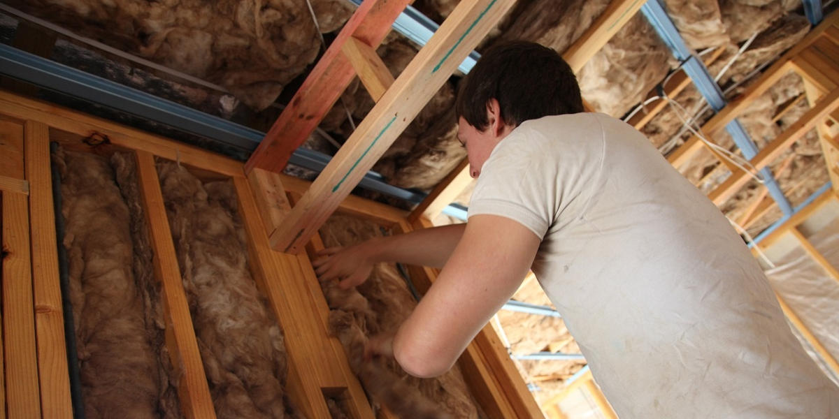 A young man in a white t-shirt in the process of installing wool insulation between wooden beams in a roof space