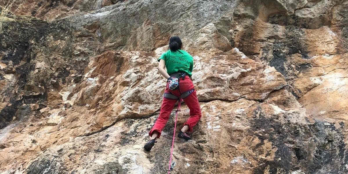 A person in red trousers and green top scales a high rock face outdoors