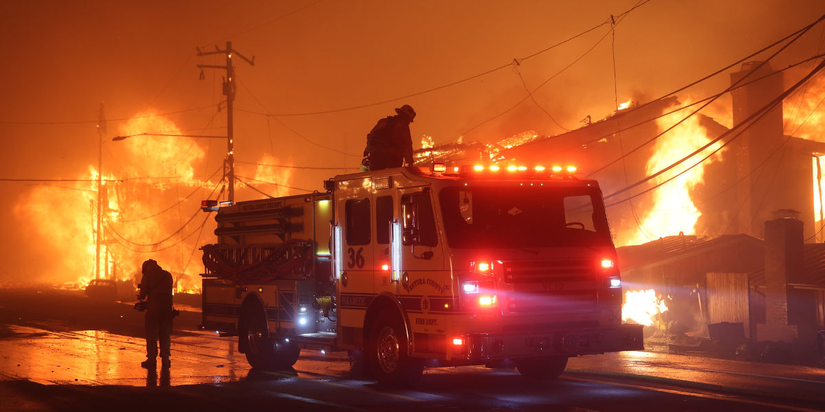 A fire engine with fire fighters along side and on top of it in the middle of a road, with burning buildings and bright fires behind it