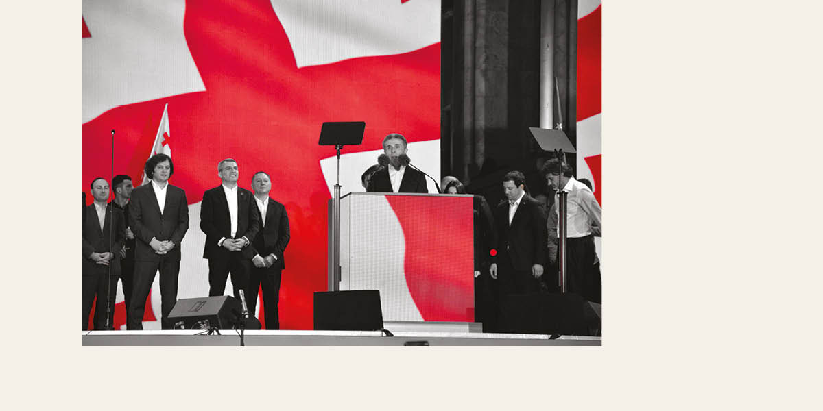 A man in a suit stands on stage behind a podium, with other men in suits around him. A huge Georgian flag provides the backdrop