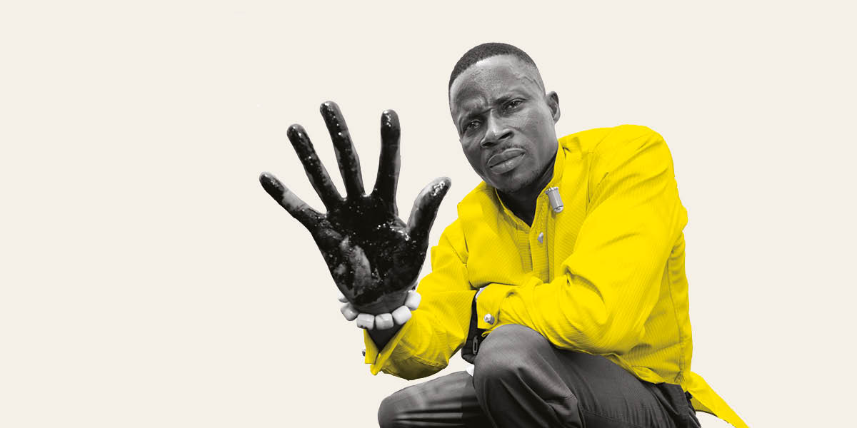 A Nigerian man in a yellow shirt holds up his hand, covered in crude oil, while looking seriously into the camera