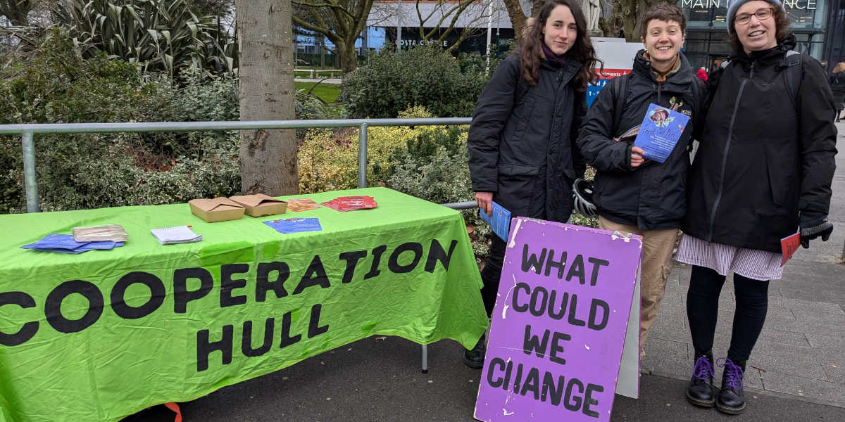 Three people standing next to a table decked in a cover reading Cooperation Hull and standing next to a sign reading 'what could we change'