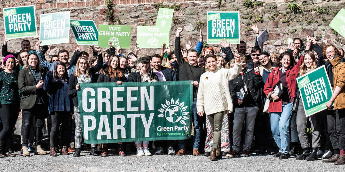 A large group of people stand behind a large Green Party banner with their fists raised above smiling faces