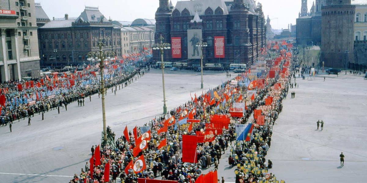 A large procession of people parading through an open square carrying flags and banners, the majority of them red